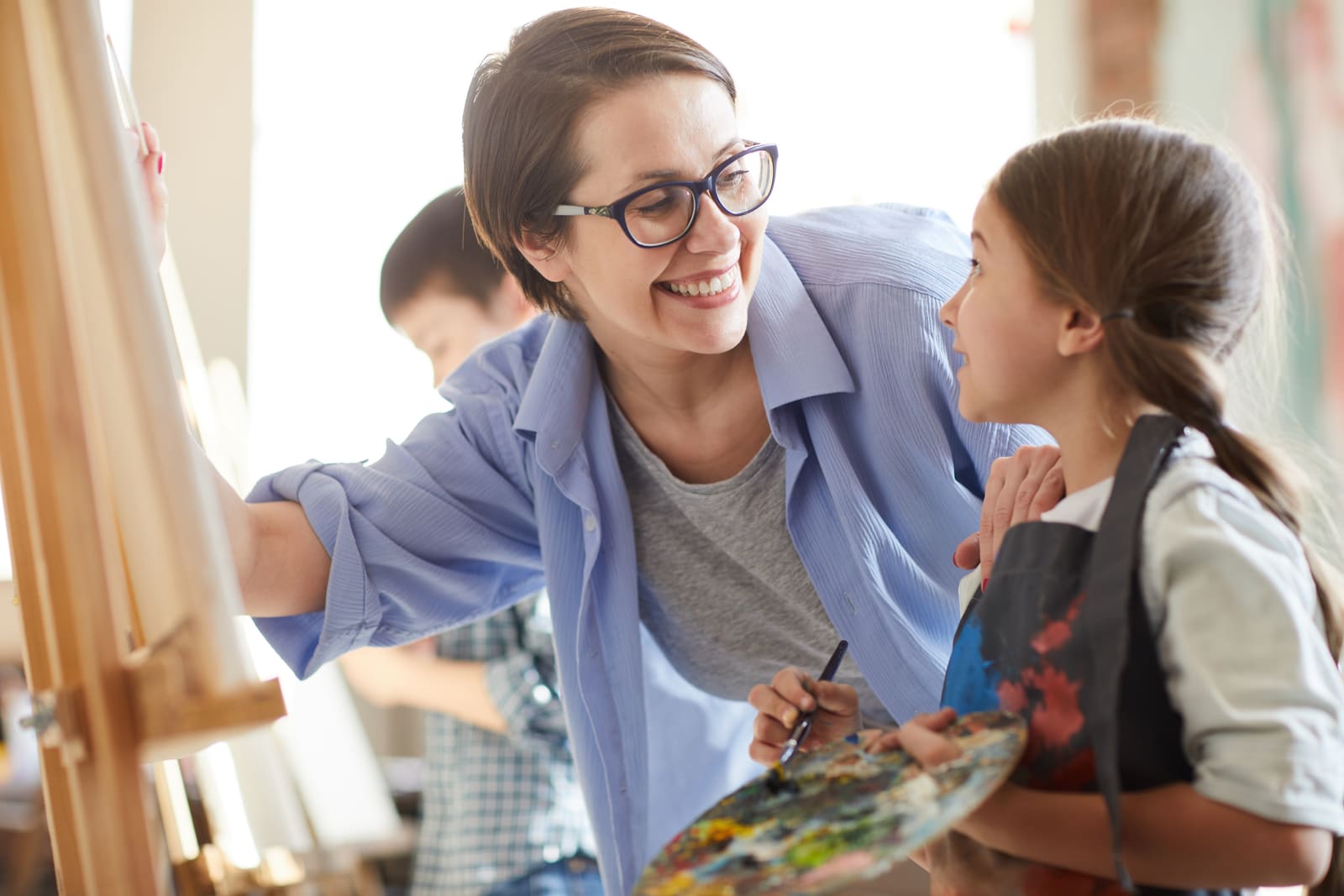 An art teacher helping a student at an easel in a bright studio classroom